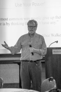 A black and white photograph of Tom Blatner speaking at a presentation. He wears a button-down checkered shirt and slacks, holding a microphone in one hand while gesturing with the other. Behind him is a projection screen displaying a title that reads "Use Your Power!" and a quote by Alice Walker: "The most common way people give up their power is by thinking they don't have any." To his right is a wooden podium.
